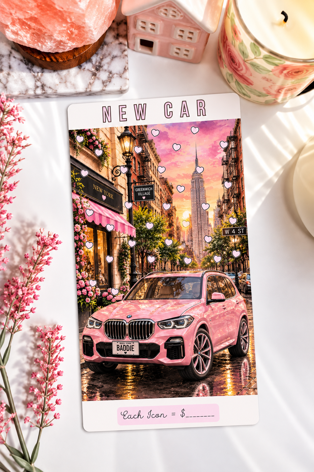 A printed card or sticker sheet featuring a pink car on a city street at sunset, surrounded by decorative props including flowers, a candle, and small house figurines on a white surface.