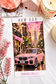 A printed card or sticker sheet featuring a pink car on a city street at sunset, surrounded by decorative props including flowers, a candle, and small house figurines on a white surface.
