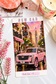A rectangular printed card or poster featuring a pink SUV on a city street with the words “NEW CAR” at the top and a handwritten note at the bottom. The card is photographed on a white surface with decorative props including pink flowers, a candle, a small house figurine, and a pink stone.