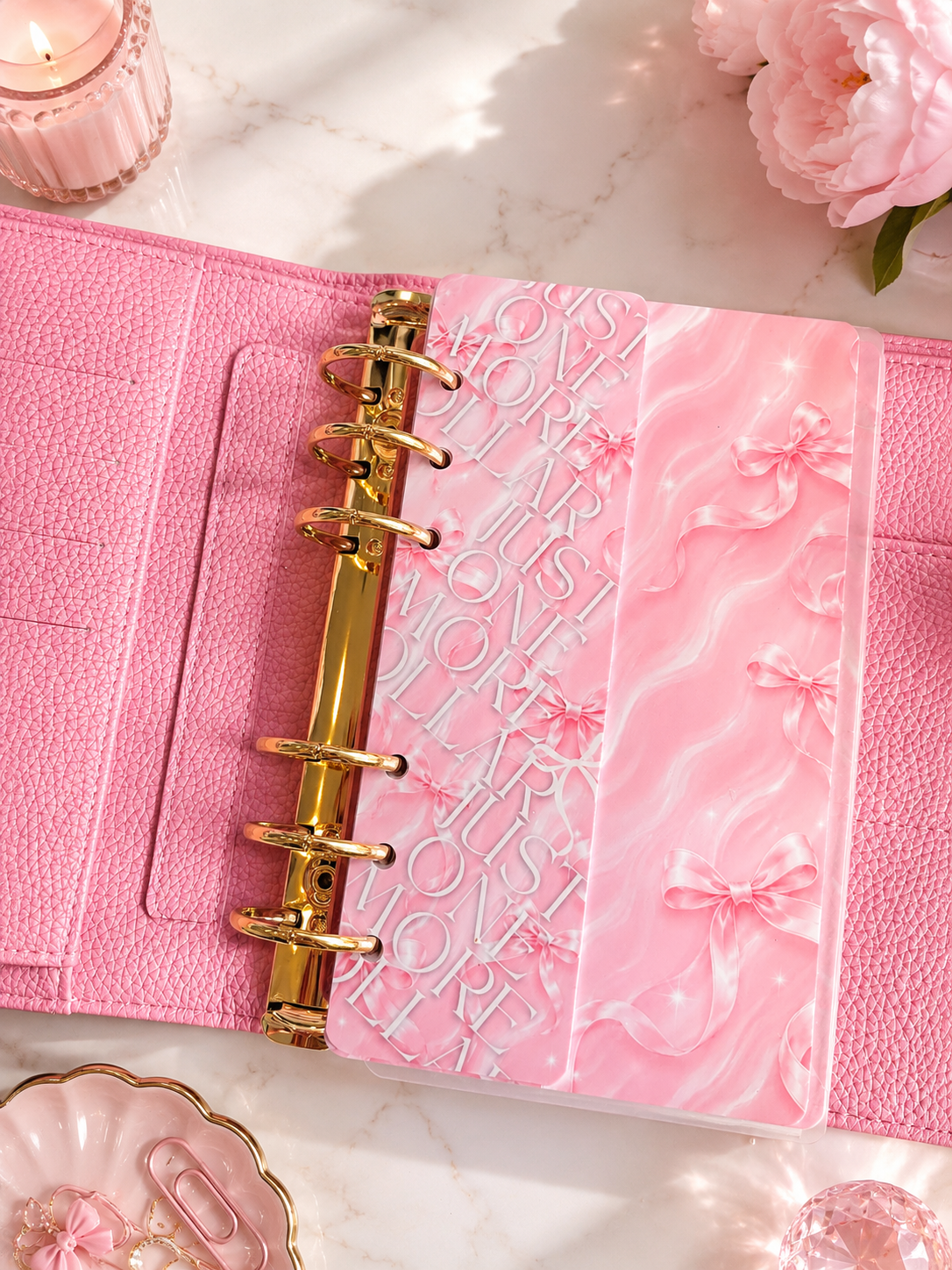 A pink ring binder planner or notebook cover laid open on a light marble surface, with embossed decorative lettering and bow motifs on the inner pages. The cover has a textured pink finish, gold-tone ring binder hardware, and is styled with pink flowers and small decorative props.