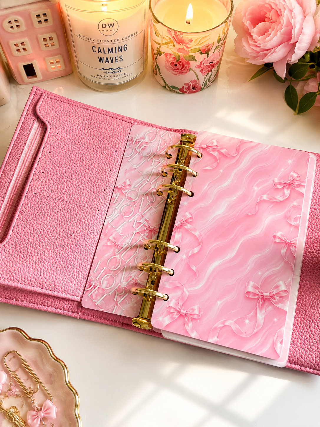 A pink textured planner binder with a marbled pink cover and gold rings, styled on a white surface with candles, a pink rose, and small decorative accessories.