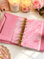 A pink textured planner binder with a marbled pink cover and gold rings, styled on a white surface with candles, a pink rose, and small decorative accessories.