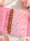A pink ring binder planner or notebook cover laid open on a light marble surface, with embossed decorative lettering and bow motifs on the inner pages. The cover has a textured pink finish, gold-tone ring binder hardware, and is styled with pink flowers and small decorative props.