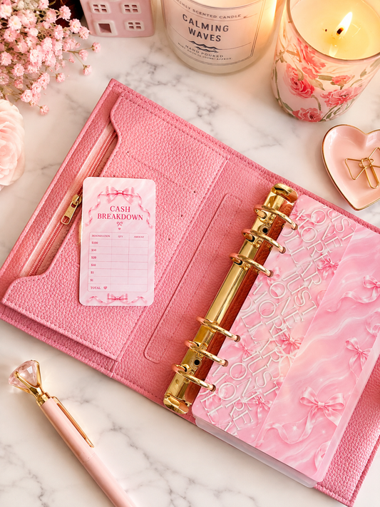 Dreamy Ribbon Cash Breakdown Card displayed inside a pink A6 cash stuffing binder with gold rings on a white marble desk, featuring a feminine pink ribbon design for tracking cash denominations.