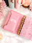 Dreamy Ribbon Cash Breakdown Card displayed inside a pink A6 cash stuffing binder with gold rings on a white marble desk, featuring a feminine pink ribbon design for tracking cash denominations.
