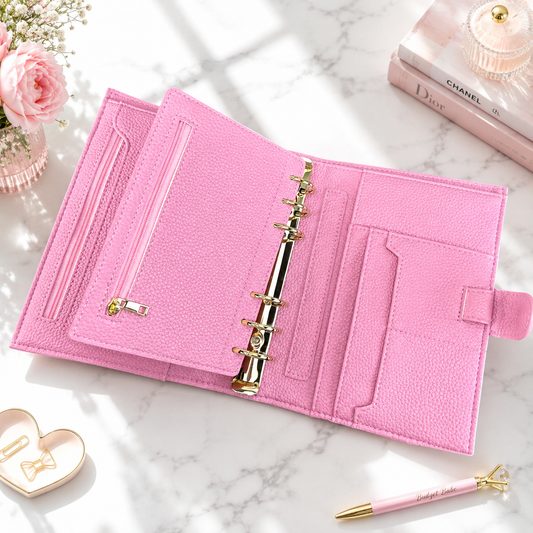 A pink textured ring binder planner or organizer shown open on a light marble surface, with card slots, pockets, and gold-tone rings. Decorative props including flowers, a pen, a small heart-shaped accessory, and stacked books are arranged around it.
