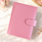 Pink A6 budget binder with cash stuffing compartments, gold rings, and zip pocket displayed on a white marble desk with lit candles and feminine pink decor.