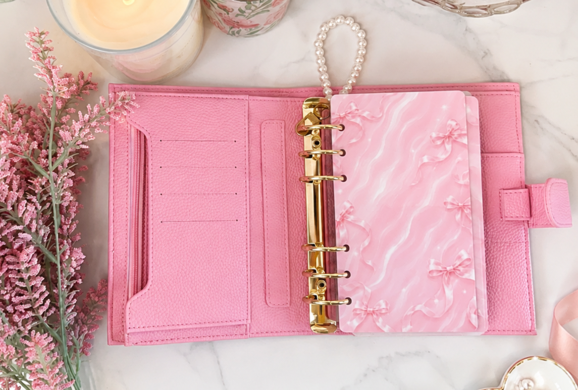 Luxury pink A6 cash stuffing binder featuring gold six-ring hardware, card slots, zippered pocket, and budgeting organizer styled on a marble desktop with aesthetic pink accessories.