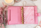Luxury pink A6 cash stuffing binder featuring gold six-ring hardware, card slots, zippered pocket, and budgeting organizer styled on a marble desktop with aesthetic pink accessories.