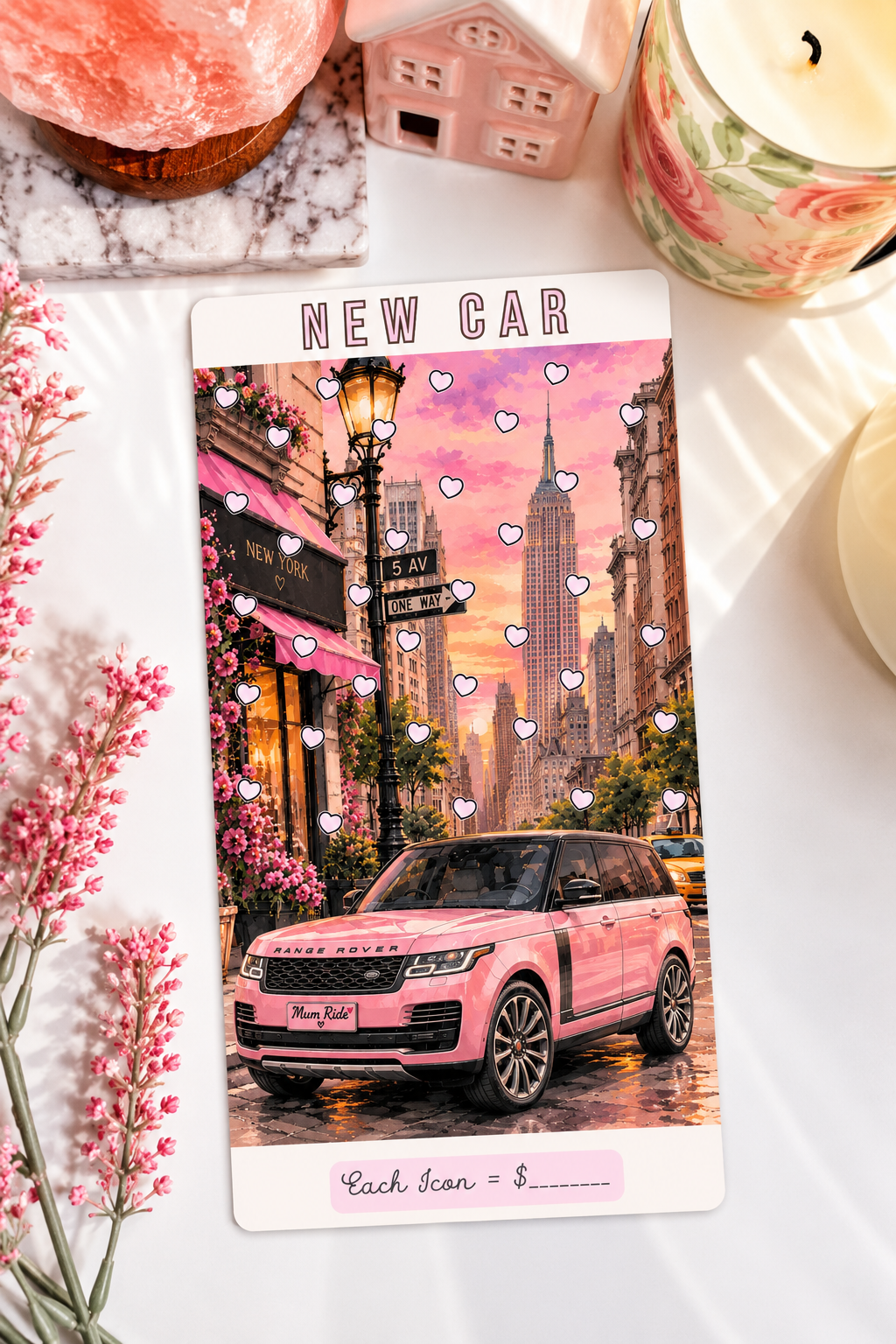 A rectangular printed card or ticket featuring a pink SUV in a city street scene with overlaid hearts and the text 'NEW CAR' at the top and 'Cash Scan = $____' at the bottom. The card is photographed on a white surface with decorative props including pink flowers, a candle, a small house figurine, and a pink stone.