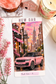 A rectangular printed card or ticket featuring a pink SUV in a city street scene with overlaid hearts and the text 'NEW CAR' at the top and 'Cash Scan = $____' at the bottom. The card is photographed on a white surface with decorative props including pink flowers, a candle, a small house figurine, and a pink stone.